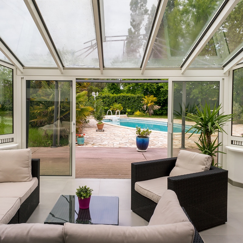Salon de véranda moderne avec toit vitré, canapés en osier foncé et vue imprenable sur la piscine et le jardin tropical aménagé.