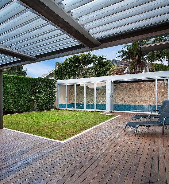 Pergola moderne et terrasse en bois pour jardin luxueux. Terrasse en bois sous une pergola bioclimatique. Le jardin verdoyant borde une piscine intérieure vitrée. Deux transats.